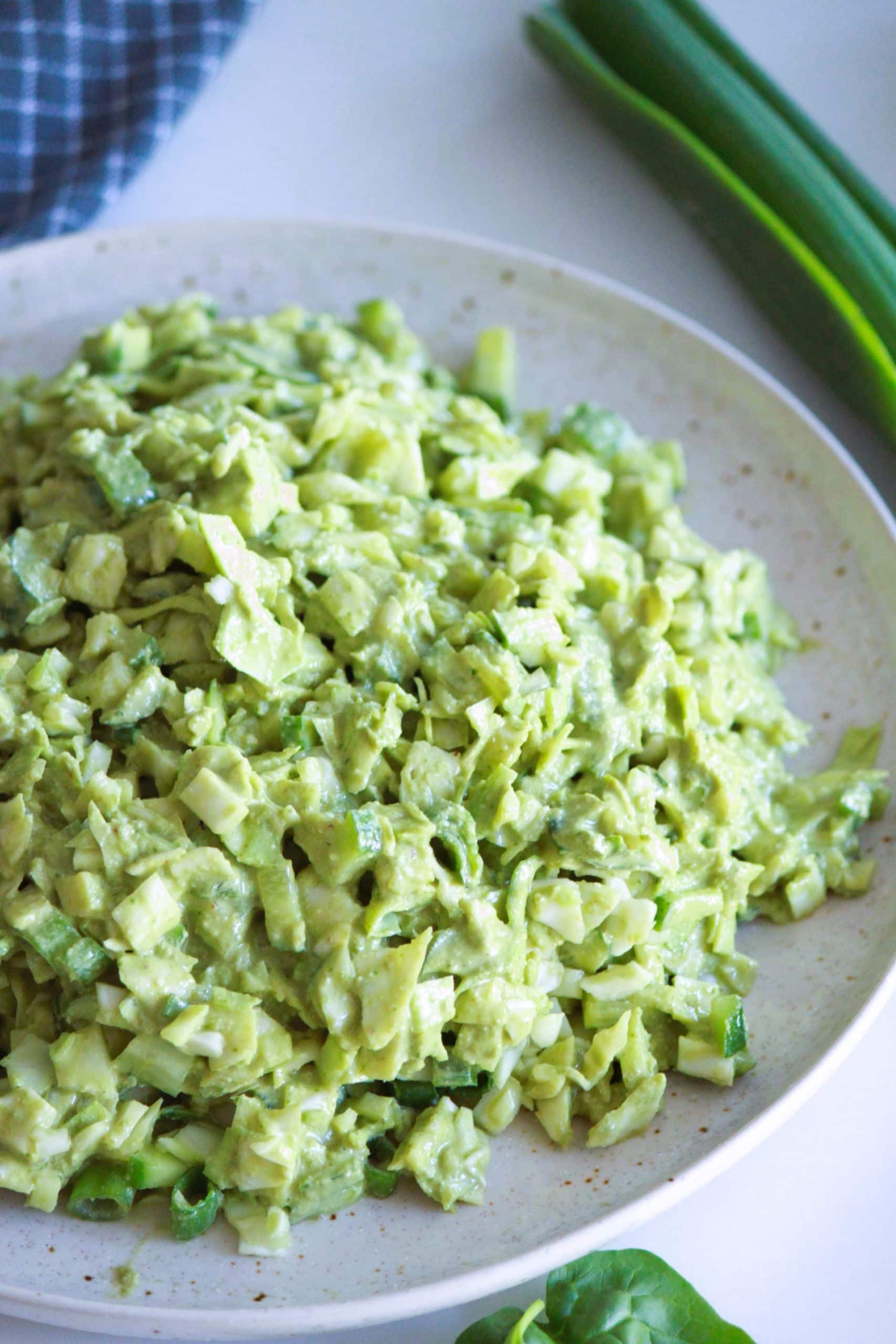 A plate of creamy avocado salad with finely chopped green vegetables, including cucumber and spring onions. Fresh green onions are visible in the background on a white surface.