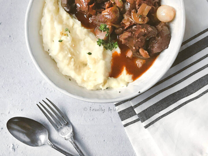 A bowl of mashed potatoes topped with beef stew, garnished with herbs, is set on a gray surface. Beside it, a striped napkin holds a fork and spoon. The dish includes chunks of beef and vegetables in a rich sauce.