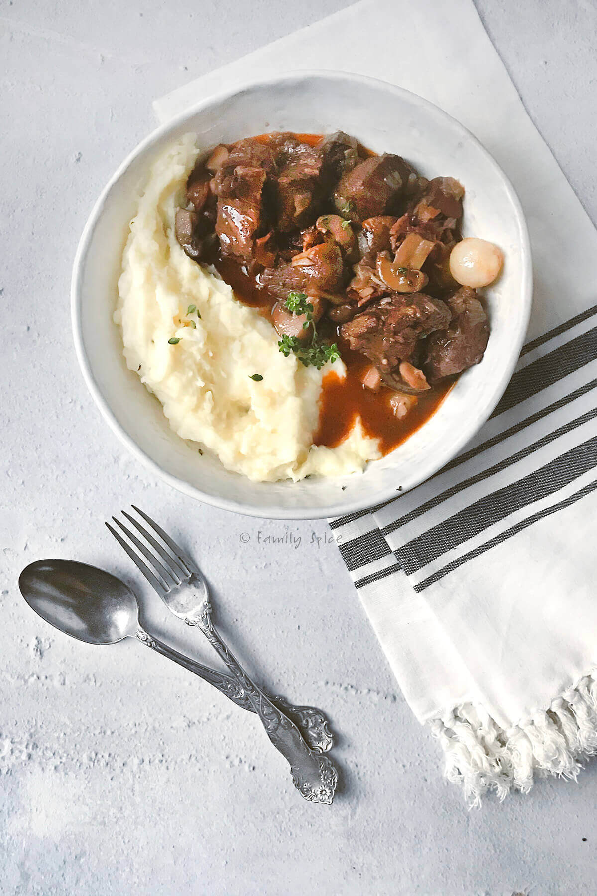 A bowl of mashed potatoes topped with beef stew, garnished with herbs, is set on a gray surface. Beside it, a striped napkin holds a fork and spoon. The dish includes chunks of beef and vegetables in a rich sauce.