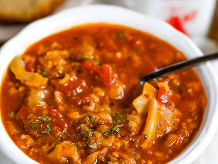 A white bowl filled with hearty cabbage roll soup, featuring chunks of cabbage, ground meat, and tomatoes. A spoon is resting in the soup. In the background, there is a slice of bread and a black-topped condiment bottle.