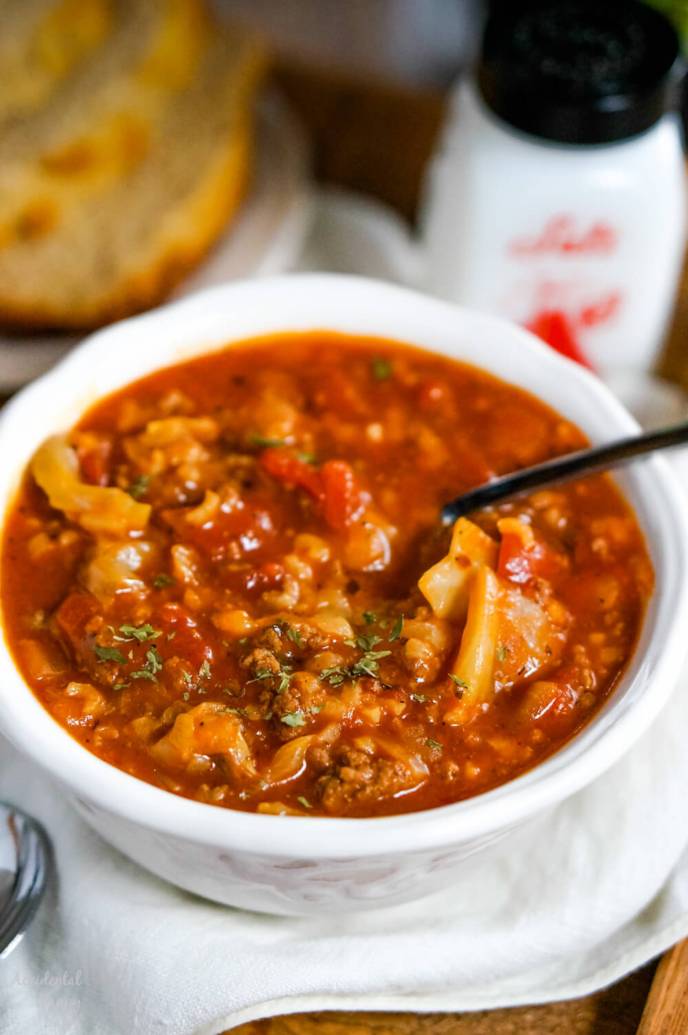 A white bowl filled with hearty cabbage roll soup, featuring chunks of cabbage, ground meat, and tomatoes. A spoon is resting in the soup. In the background, there is a slice of bread and a black-topped condiment bottle.