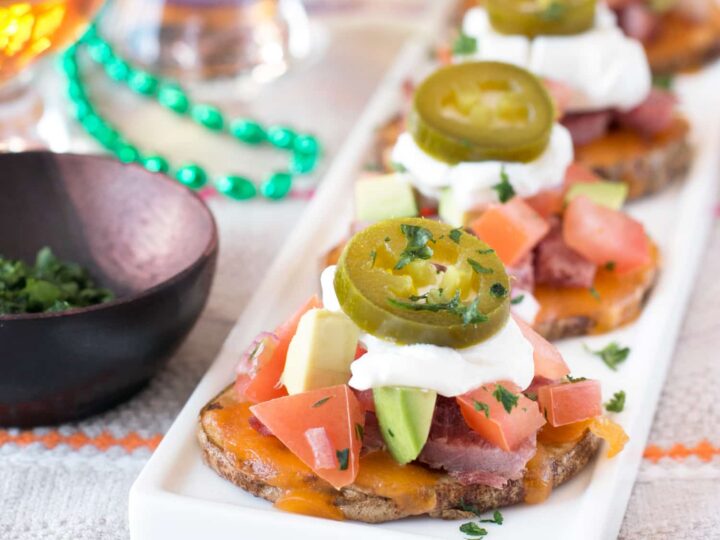 Close-up of loaded potato slices on a white platter. Each slice is topped with melted cheese, diced tomatoes, avocado, sour cream, jalapeño slices, and herbs. A bowl of herbs and two glasses of amber liquid in the background.