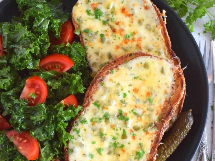 A plate with two slices of bread topped with melted cheese, labeled Irish Gherkin Rarebit. The dish is garnished with herbs and served alongside a salad with kale and tomatoes, and pickles.