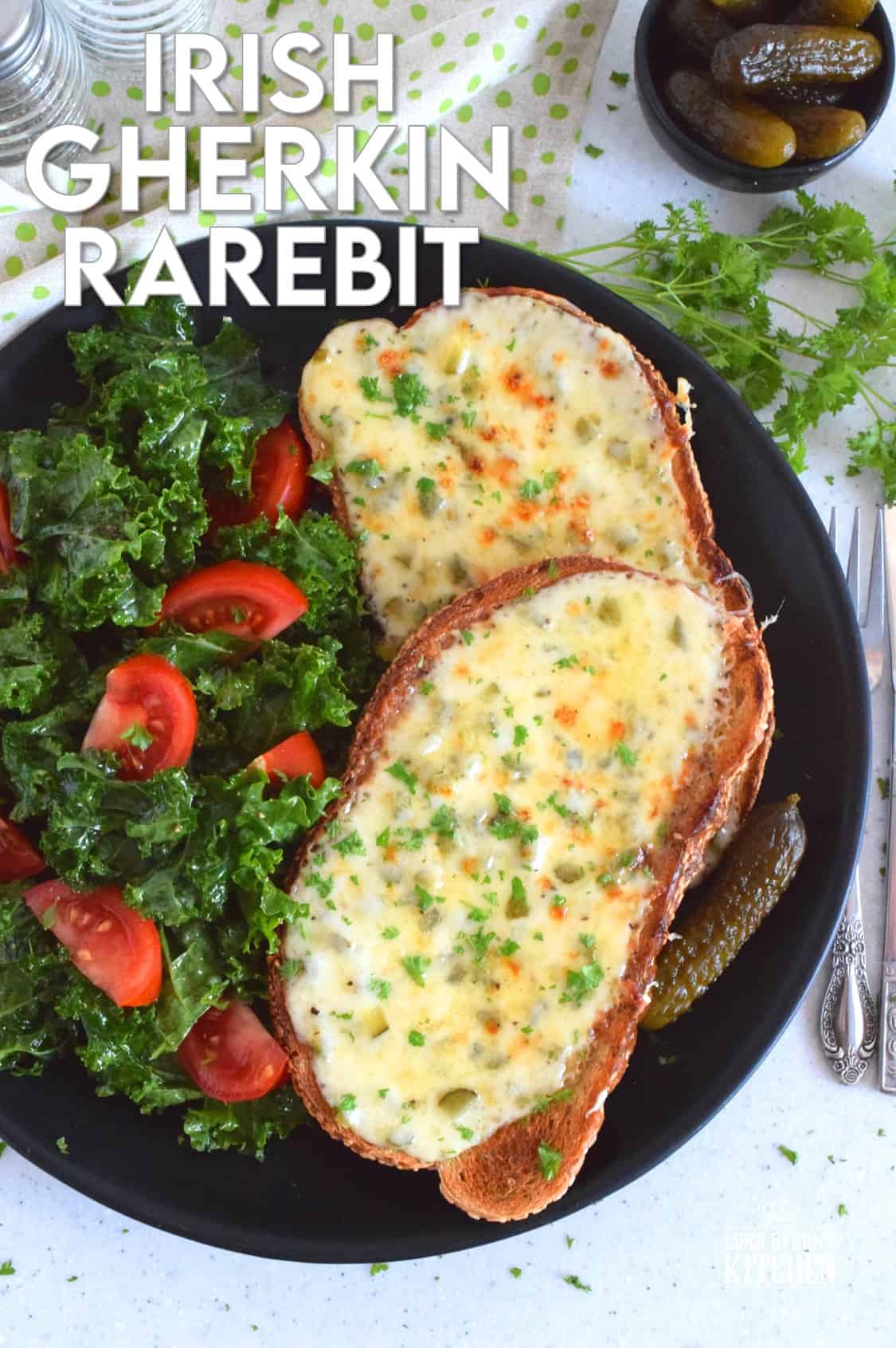 A plate with two slices of bread topped with melted cheese, labeled Irish Gherkin Rarebit. The dish is garnished with herbs and served alongside a salad with kale and tomatoes, and pickles.