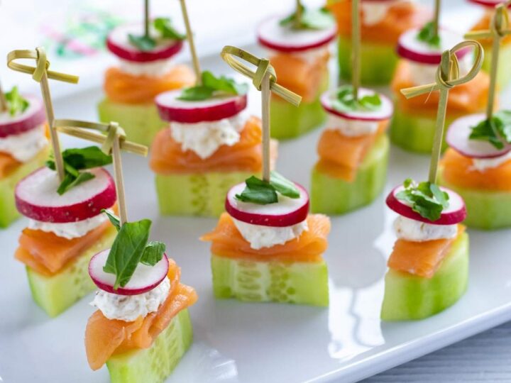Cucumber slices topped with smoked salmon, a dollop of cream cheese, a radish slice, and a sprig of greens, all skewered with decorative picks, are arranged on a white platter.