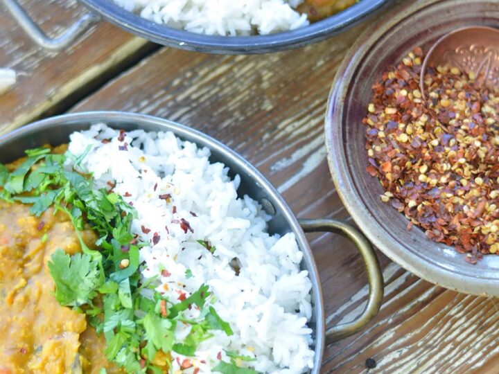 Two bowls of curry and rice are garnished with cilantro and chili flakes. A small dish of chili flakes is nearby. The bowls rest on a rustic wooden surface.