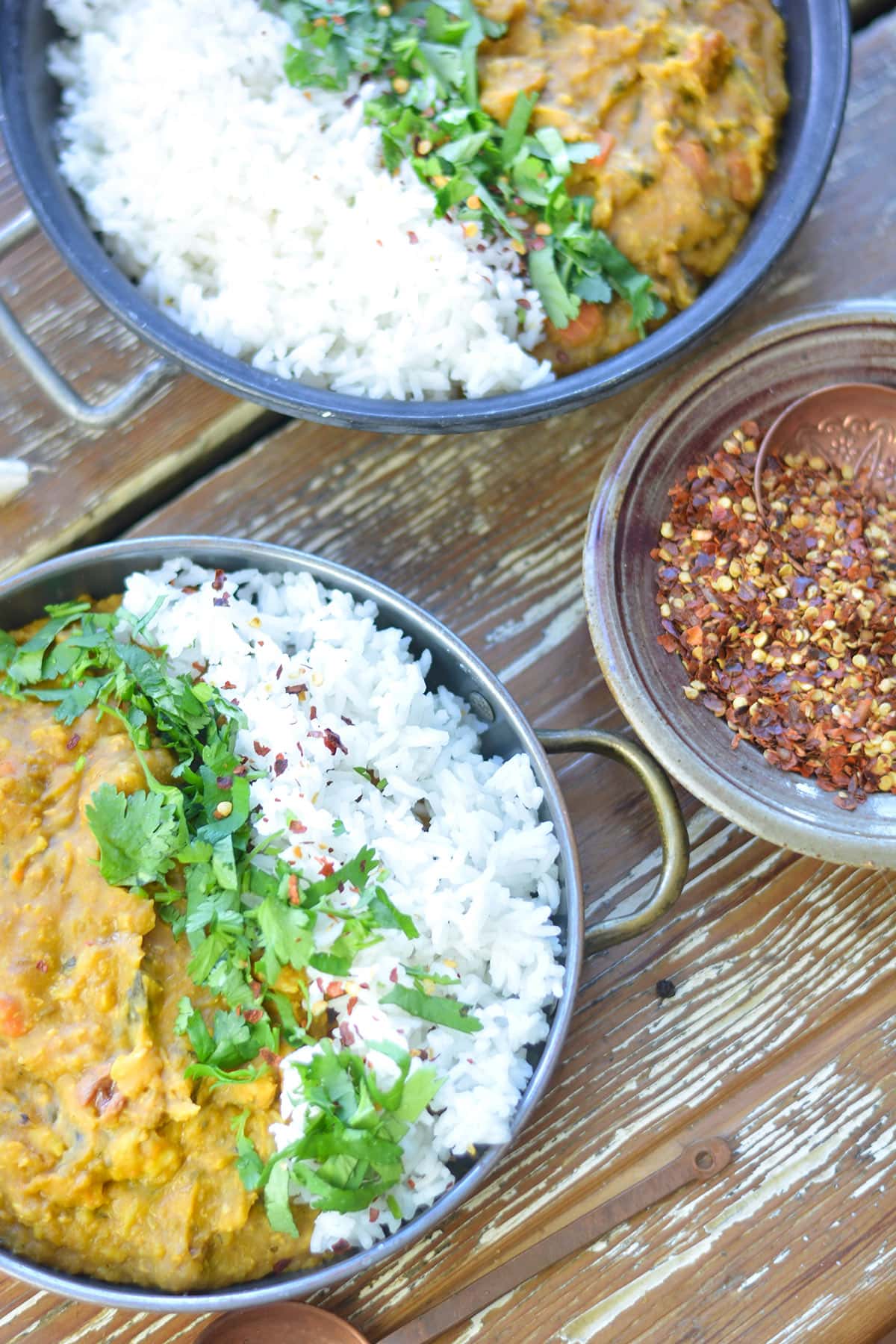 Two bowls of curry and rice are garnished with cilantro and chili flakes. A small dish of chili flakes is nearby. The bowls rest on a rustic wooden surface.