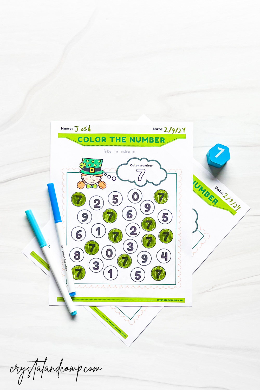 The image shows a worksheet titled Color the Number with a frog illustration at the top. The number 7 is highlighted. Several circles with numbers are marked with a green crayon. Two markers and a blue dice with the number 7 are beside the sheet.