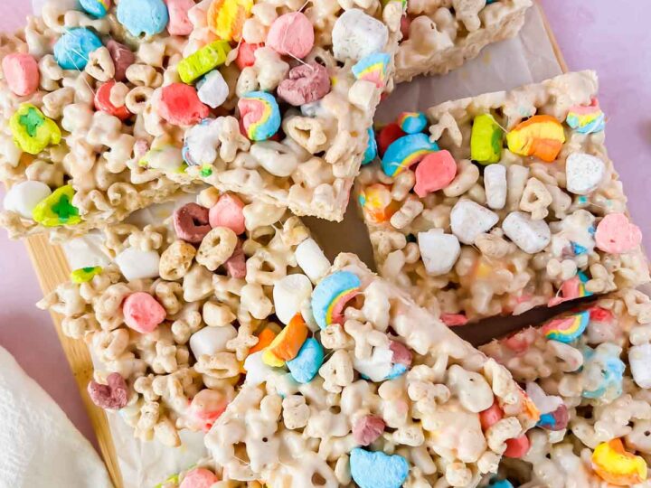 A wooden board with cereal treats, featuring colorful marshmallows and oat rings, cut into square pieces. The background is a light pink surface with a white cloth partially visible.