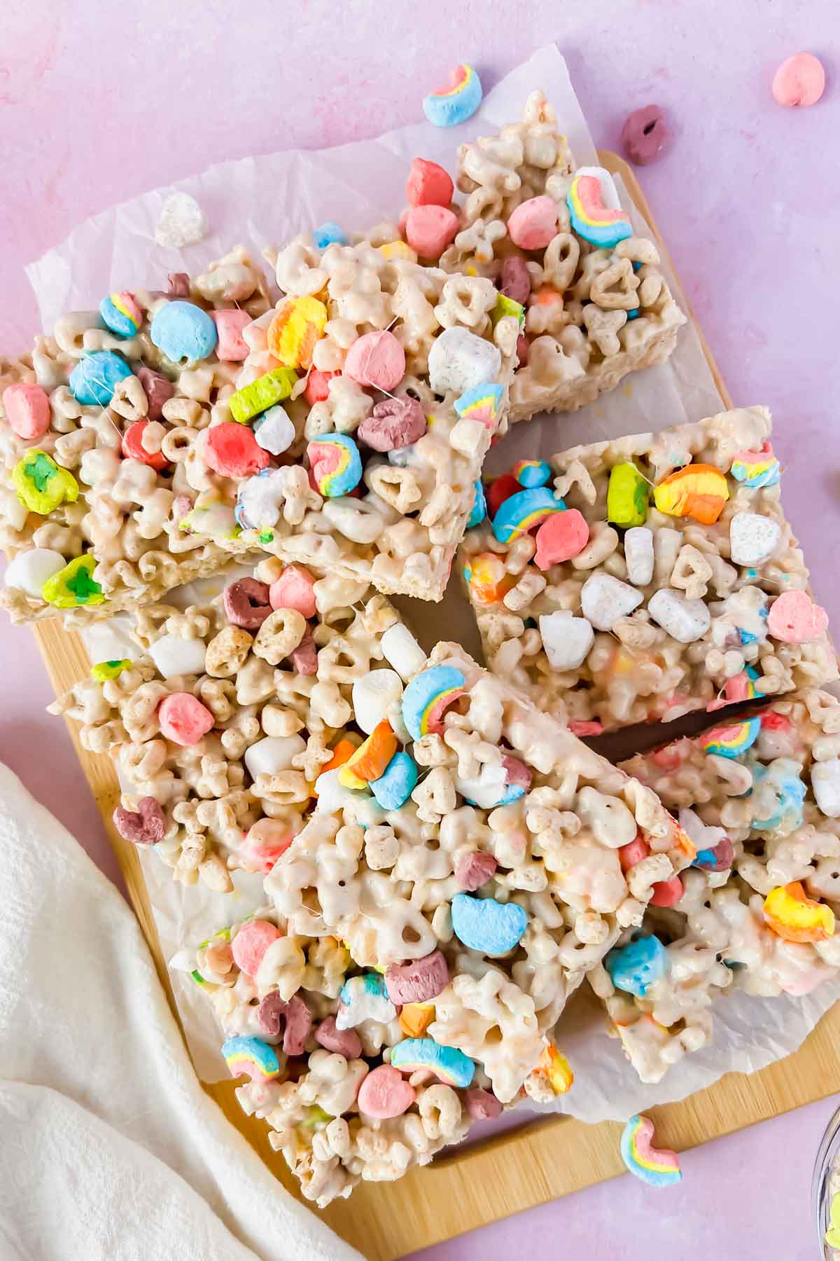 A wooden board with cereal treats, featuring colorful marshmallows and oat rings, cut into square pieces. The background is a light pink surface with a white cloth partially visible.