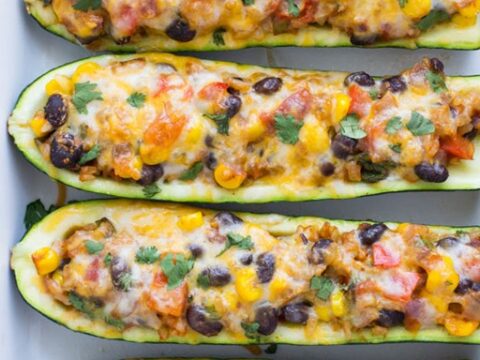 Four zucchini halves are filled with a colorful mixture of melted cheese, black beans, corn, and diced tomatoes, garnished with cilantro, on a white platter.
