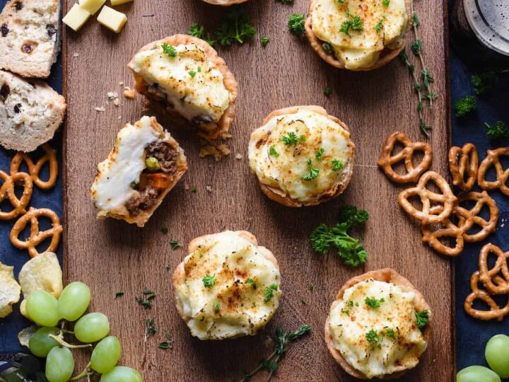 A wooden board features baked potatoes filled with creamy topping, surrounded by grapes, cheese cubes, sliced bread, pretzels, and chips. Sprigs of herbs garnish the scene. Glasses with drinks are partially visible at the edges.