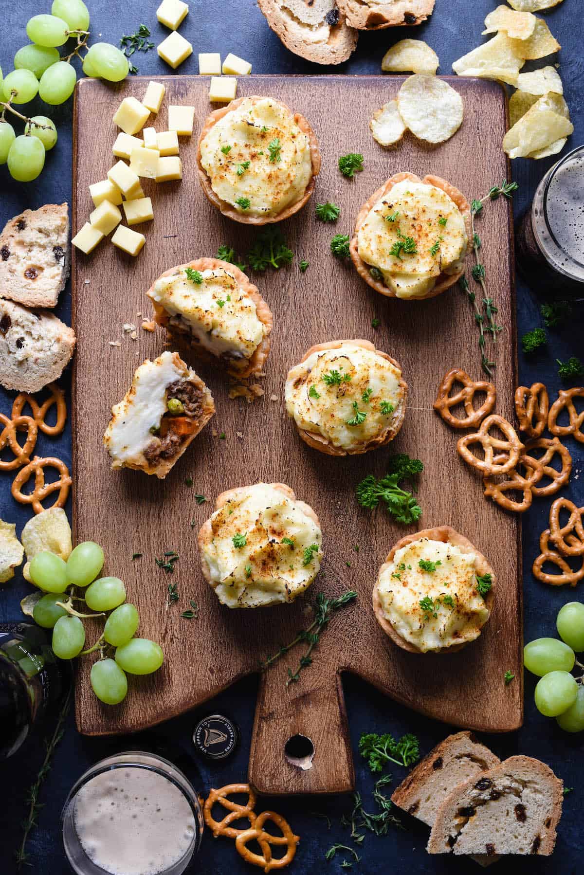 A wooden board features baked potatoes filled with creamy topping, surrounded by grapes, cheese cubes, sliced bread, pretzels, and chips. Sprigs of herbs garnish the scene. Glasses with drinks are partially visible at the edges.