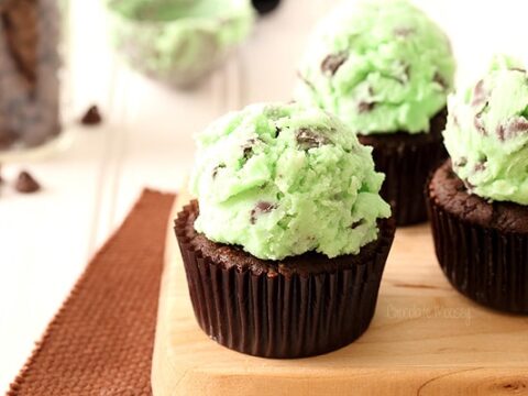 Three chocolate cupcakes topped with scoops of mint chocolate chip frosting sit on a wooden board. The frosting is pale green with chocolate chunks. A textured brown mat and a jar of chocolate chips are in the background.
