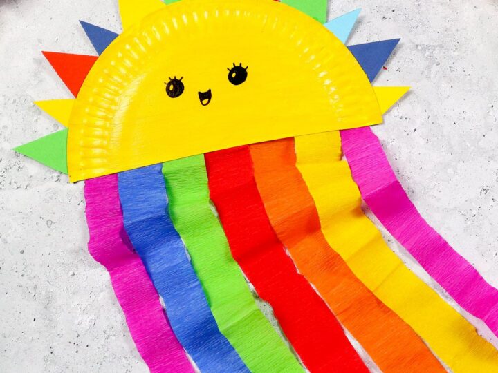 A colorful craft sun made from a yellow painted paper plate with cute eyes and a smile. Triangular sun rays in various colors stick out from the top. Multicolored crepe paper streamers hang below, resembling rainbow rays.
