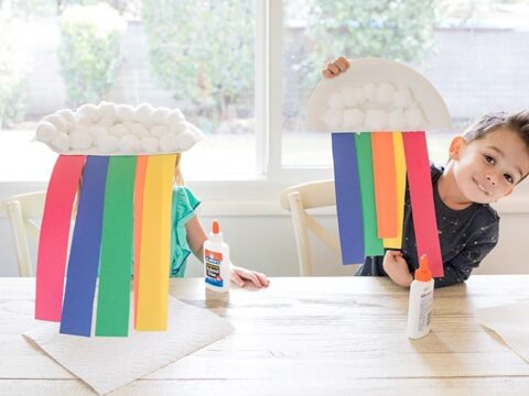 Two children sit at a table with rainbow crafts made from paper plates, cotton balls, and colorful paper strips. Glue bottles are on the table. The window in the background reveals a garden view.