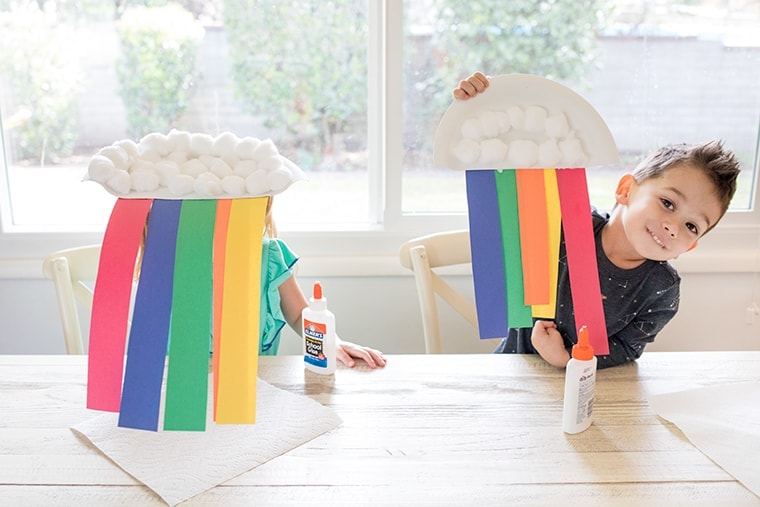 Two children sit at a table with rainbow crafts made from paper plates, cotton balls, and colorful paper strips. Glue bottles are on the table. The window in the background reveals a garden view.