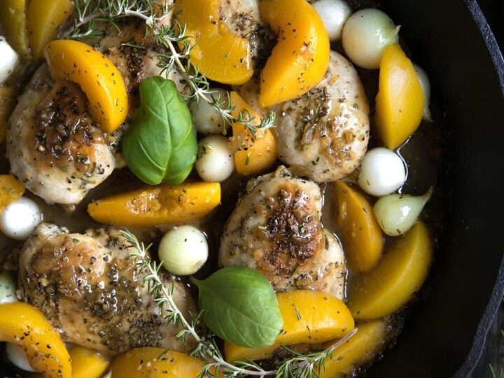 A skillet with cooked chicken breasts, bright yellow peach slices, pearl onions, and fresh green basil leaves, garnished with sprigs of rosemary. The dish rests on a dark wooden surface.