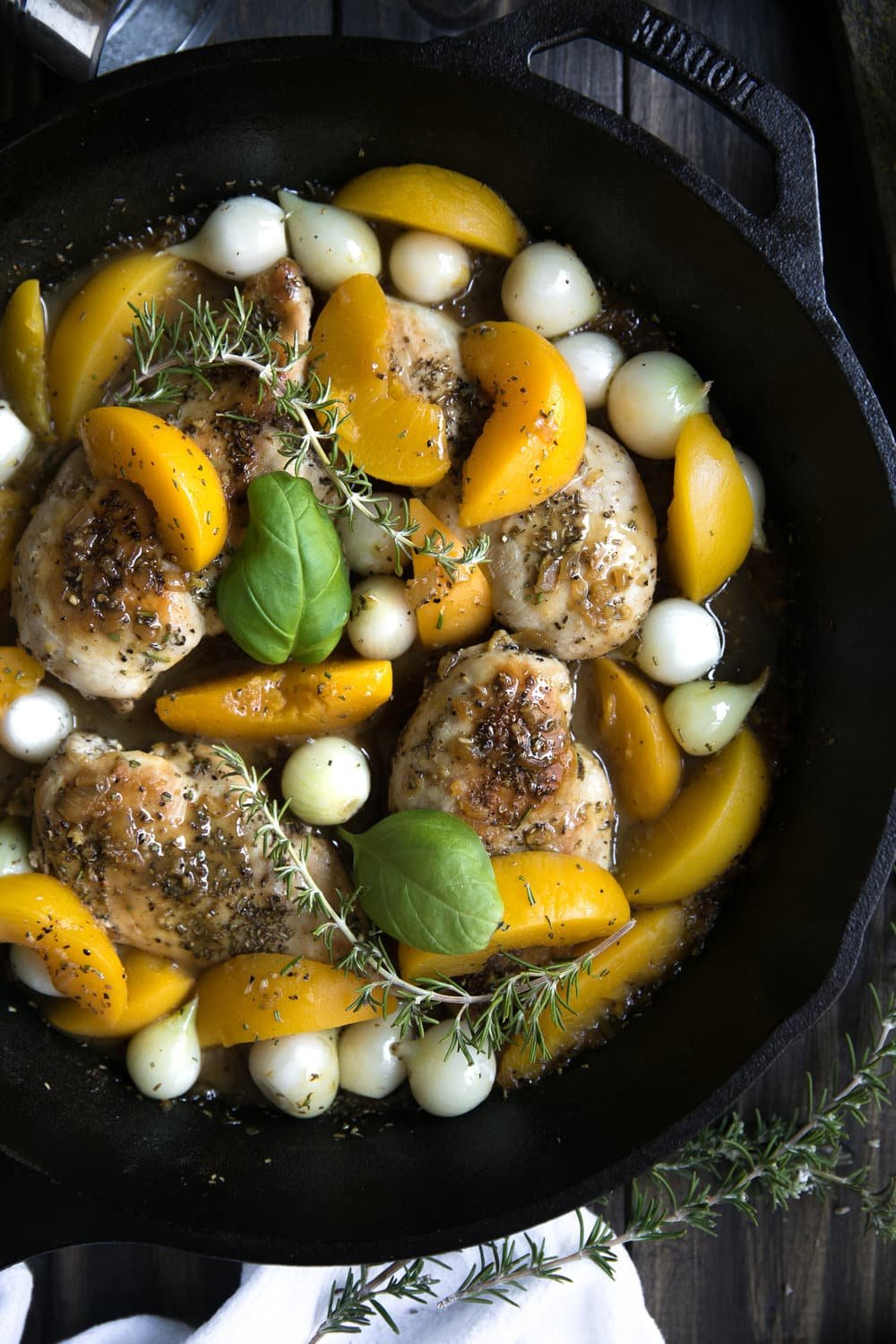A skillet with cooked chicken breasts, bright yellow peach slices, pearl onions, and fresh green basil leaves, garnished with sprigs of rosemary. The dish rests on a dark wooden surface.