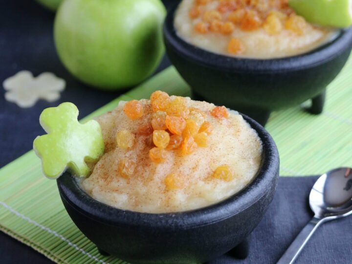 Two black bowls of applesauce topped with golden raisins are garnished with apple slices shaped like clovers. They are set on a green napkin with whole green apples in the background and a spoon beside them.