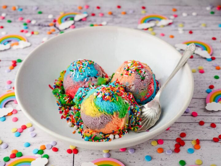 A white bowl holds three scoops of colorful rainbow ice cream topped with sprinkles. A spoon rests inside the bowl. The table is scattered with rainbow decorations and confetti.