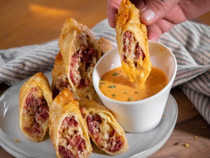 A plate of sliced crispy egg rolls filled with meat and cheese, next to a small white bowl of orange dipping sauce. A hand is dipping one of the egg rolls into the sauce. A striped cloth is in the background.