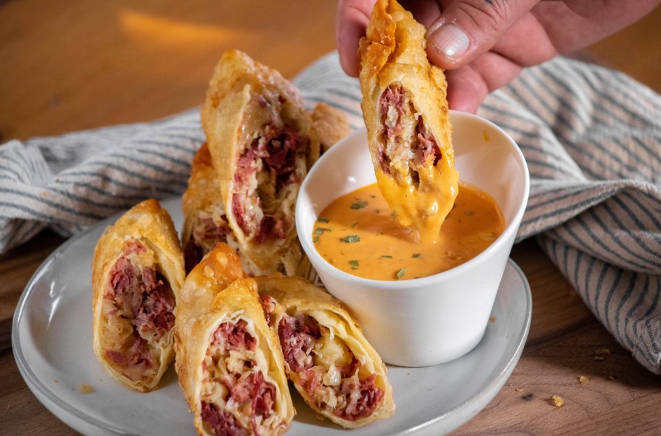 A plate of sliced crispy egg rolls filled with meat and cheese, next to a small white bowl of orange dipping sauce. A hand is dipping one of the egg rolls into the sauce. A striped cloth is in the background.