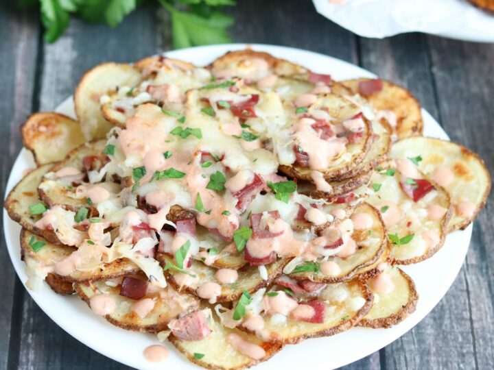 A plate of crispy potato slices topped with melted cheese, chopped red onions, parsley, and a drizzle of creamy sauce. Garnished with fresh parsley in the background on a wooden table.
