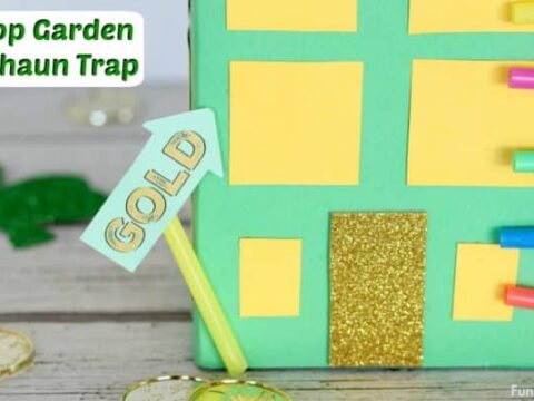 A colorful homemade leprechaun trap resembling a building with a glittery gold door and rainbow-colored straws on the side. A small sign reading GOLD and paper shamrocks and coins decorate the wooden surface.