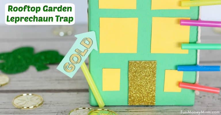 A colorful homemade leprechaun trap resembling a building with a glittery gold door and rainbow-colored straws on the side. A small sign reading GOLD and paper shamrocks and coins decorate the wooden surface.