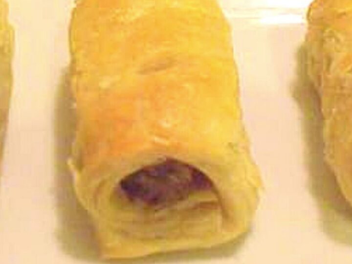 A close-up image of a puff pastry roll with a golden-brown crust, partially showing its flaky texture and the filling inside. The roll is placed on a light-colored surface.
