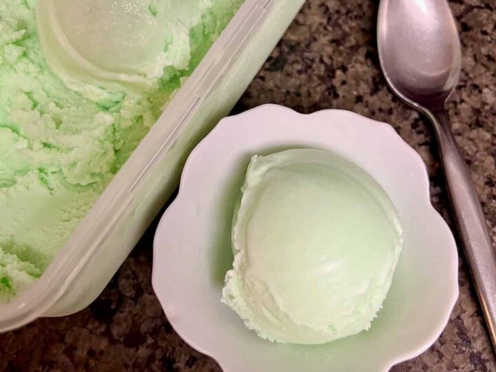 A small bowl with a scoop of light green ice cream sits on a countertop next to a full container of the same ice cream. A spoon lies beside the bowl.