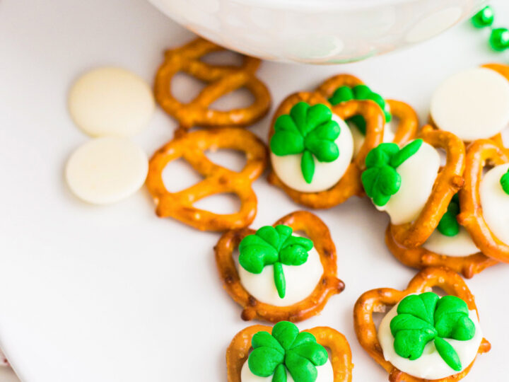 A white plate and bowl with pretzels topped with melted white chocolate and green clover-shaped decorations. Some pretzels and white chocolate discs are scattered around.