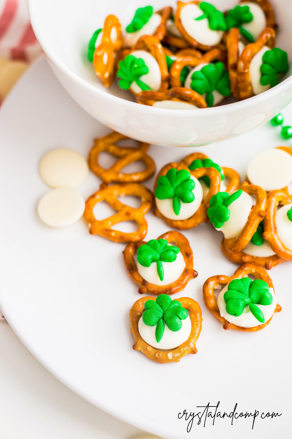 A white plate and bowl with pretzels topped with melted white chocolate and green clover-shaped decorations. Some pretzels and white chocolate discs are scattered around.