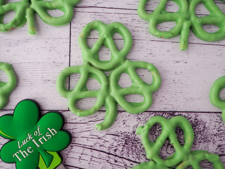 Green pretzels arranged in shamrock shapes are placed on a white wooden surface. A clover-shaped decoration with the words Luck of the Irish is also visible on the surface.