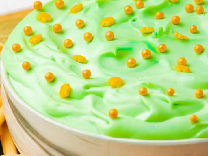 A bowl filled with light green dip, topped with small golden candy balls. The dip is surrounded by cookies and pretzels on a tray.
