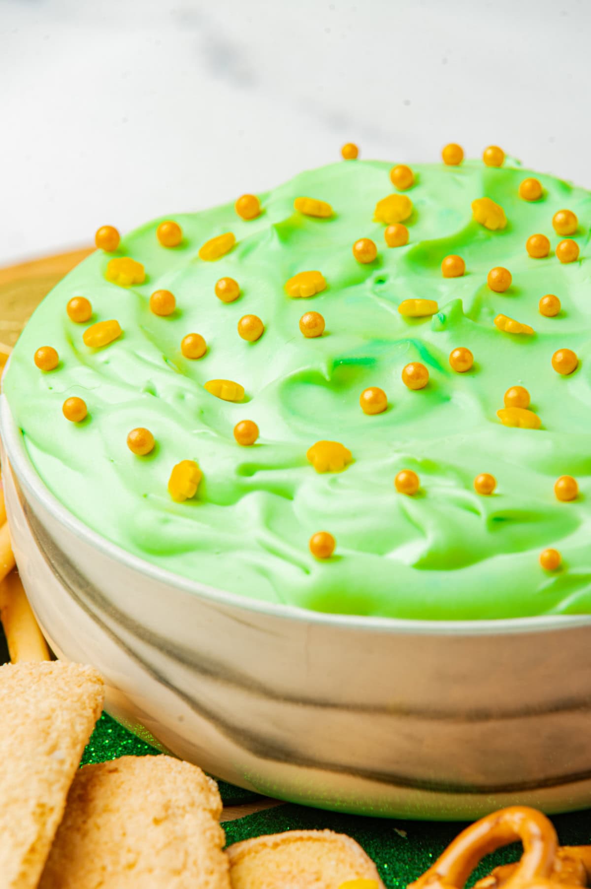 A bowl filled with light green dip, topped with small golden candy balls. The dip is surrounded by cookies and pretzels on a tray.