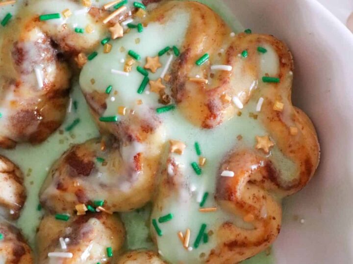 Close-up of St. Patrick’s Day-themed cinnamon rolls topped with green icing. The rolls are sprinkled with green, gold, and white sprinkles, including star-shaped ones, and are served in a white dish.