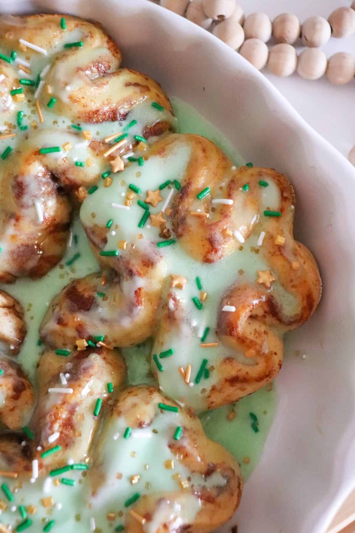 Close-up of St. Patrick’s Day-themed cinnamon rolls topped with green icing. The rolls are sprinkled with green, gold, and white sprinkles, including star-shaped ones, and are served in a white dish.
