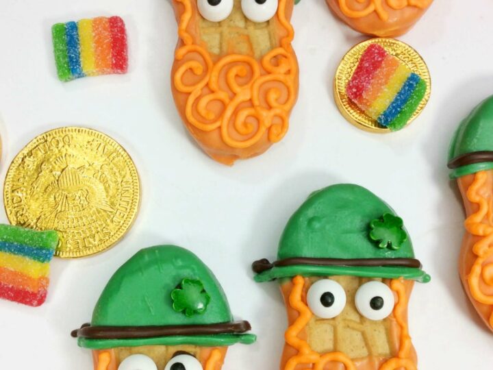 Cookies decorated as leprechauns with orange beards, green hats, and googly eyes are arranged with gold chocolate coins and rainbow candy strips on a white background. Text reads, St. Patricks Day Leprechaun Cookies.