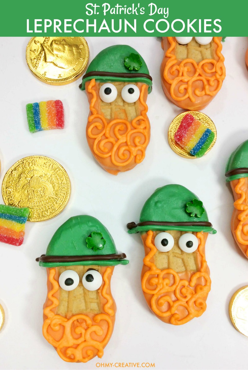 Cookies decorated as leprechauns with orange beards, green hats, and googly eyes are arranged with gold chocolate coins and rainbow candy strips on a white background. Text reads, St. Patricks Day Leprechaun Cookies.
