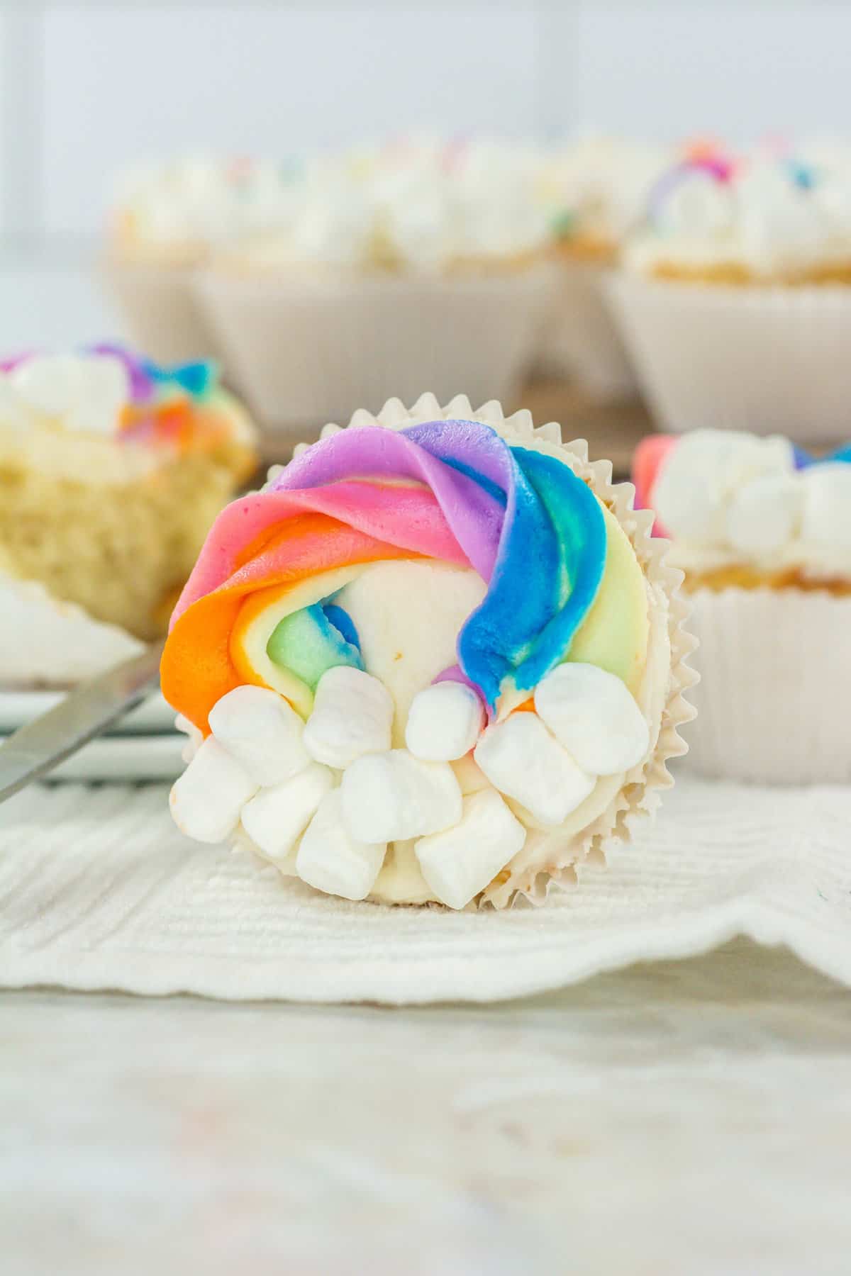 Close-up of a cupcake with vibrant, swirled frosting in pink, orange, purple, and blue, topped with mini marshmallows. The cupcake is placed on a white napkin, with more cupcakes blurred in the background.