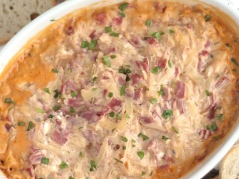 A baked Reuben dip in a white dish, topped with chopped parsley. The creamy dip is mixed with visible shredded ingredients. Slices of pumpernickel and rye bread are arranged around the dish for dipping.