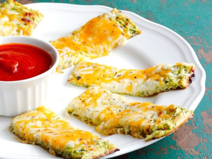 A white plate with rectangular cheesy garlic bread pieces topped with herbs and a mix of melted cheeses. A small white ramekin filled with marinara sauce is placed on the plates edge. The plate rests on a teal, rustic surface.