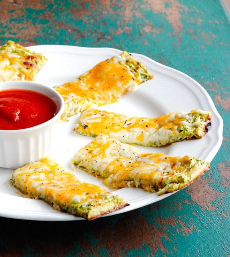 A white plate with rectangular cheesy garlic bread pieces topped with herbs and a mix of melted cheeses. A small white ramekin filled with marinara sauce is placed on the plates edge. The plate rests on a teal, rustic surface.