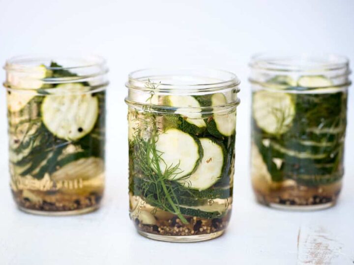 Three glass jars filled with sliced cucumbers, dill, and spices, presumably for pickling, are arranged against a light background. The jars are transparent, showing the contents clearly.