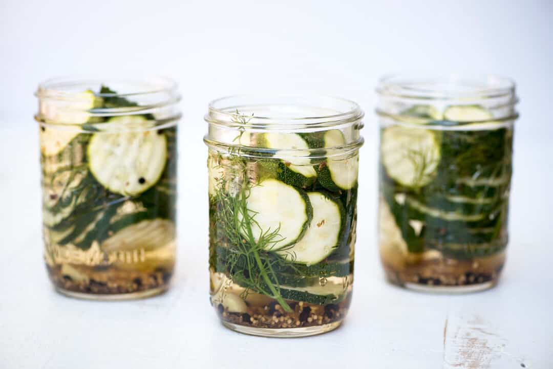 Three glass jars filled with sliced cucumbers, dill, and spices, presumably for pickling, are arranged against a light background. The jars are transparent, showing the contents clearly.