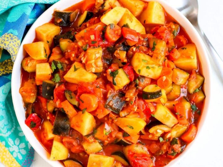 A bowl of colorful vegetable stew, featuring chunks of potato, zucchini, eggplant, and bell peppers, in a rich tomato sauce. A spoon is placed beside the bowl on a patterned blue cloth.