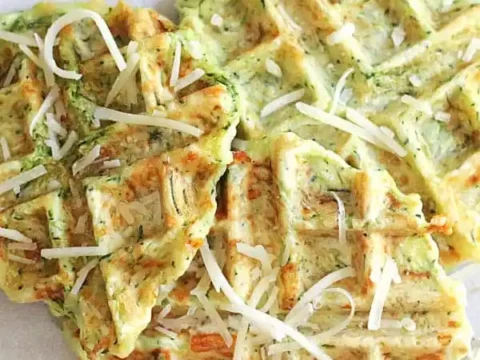 Zucchini Parmesan waffles on a white surface, topped with shredded cheese. The waffles have a golden-brown, crispy appearance. Perfect for cooking enthusiasts, this recipe shines as Zucchini Parmesan Waffles graces the top with style.