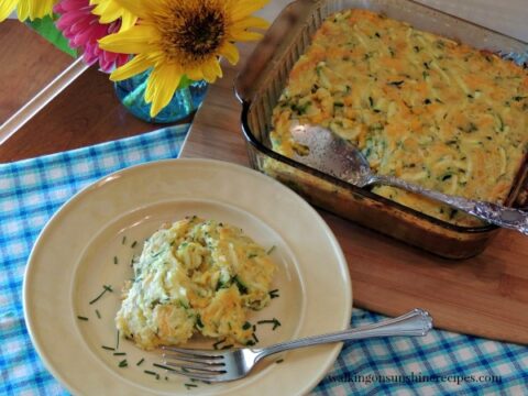 A plate with a serving of zucchini and cheese casserole, garnished with herbs. The casserole dish is next to it on a checkered blue cloth. A fork is on the plate, and yellow flowers are in the background.